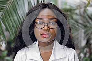 Close up portrait of young African woman, scientist, botanist, agronomist, wearing glasses and white lab coat, posing to