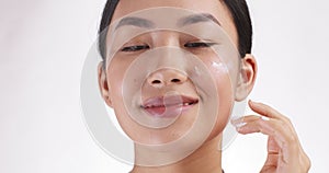 Close up portrait of young asian lady applying cream on cheek, white studio background, slow motion