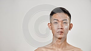 Close up portrait of young asian man with problematic skin applied cream on his face, looking at camera  over