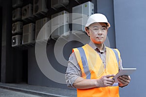 Close-up portrait of a young Asian man wearing a hard hat and vest, standing outside a factory, construction site