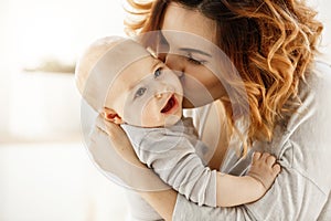 Close up portrait of young attractive mother kissing his crying baby. Kid looking aside with frightened face expression