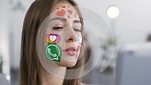 Close-up portrait of young attractive woman with painted social media icons on her face holding phone and tablet in