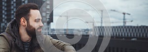 Close-up portrait of young bearded man sitting on top roof, on background of city buildings. Copy space concept.
