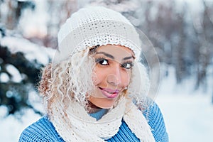 Close up portrait of young beautiful girl with afro hair wearing hat, blue sweater posing in winter park.