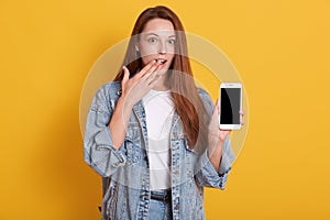 Close up portrait of young beautiful shocked woman dresses casual style, holding phone with blank screen and keeps palm on her