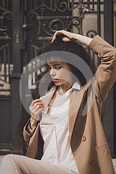 Close up portrait of young beautiful woman with long brunette curly hair posing against building background. sitting on