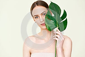 Close up portrait of young beautiful woman with perfect smooth skin covering her eye with monstera tropical leaf