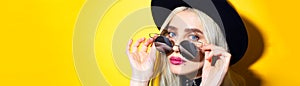 Close-up portrait of young blonde girl, wearing black hat and choker, taking off sunglasses on yellow background with copy space.