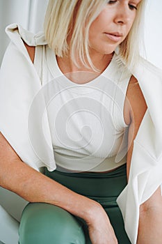 Close up portrait of young blonde woman in sportswear sitting on background of white wall