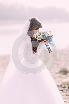 Close-up portrait of young bride in white dress and veil sniffing wedding bouquet with blue bow outdoors