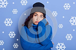 Close up portrait of young brunette female standing over studio wall with snowflakes on background, keep her hand under chin and