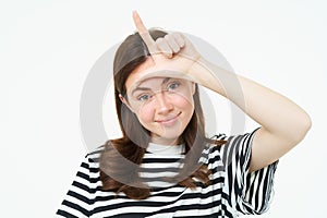 Close up portrait of young brunette girl mocking you, showing loser finger gesture, l letter with hand on forehead