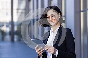 Close-up portrait of a young business woman standing outside an office building, holding a tablet and smiling at the