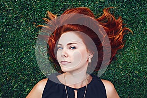 Close up portrait of young Caucasian beautiful girl woman with red brown hair lying on green grass in park. View from above