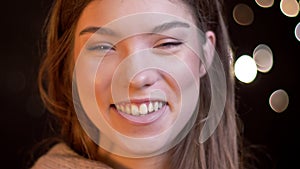 Close-up portrait of young caucasian girl laughing shyly into camera on blurred lights background.