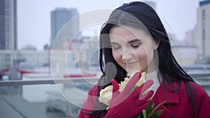 Close-up portrait of young Caucasian woman with black hair smelling yellow roses and smiling. Charming lady standing