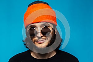 Close-up portrait of young cheerful guy looking up, wearing orange hat and sunglasses on blue background.