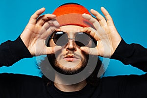 Close-up portrait of young confident man, holding hands on sunglasses that he wear. Wearing orange hat and black sweater. Studio
