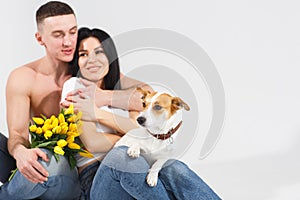 Close up portrait young couple sit and hugging, holding yellow flowers and dog in studio on white background. couple