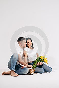 Close up portrait young couple sit and hugging, holding yellow flowers and dog in studio on white background. couple