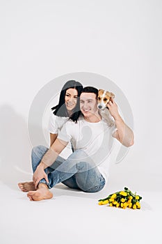 Close up portrait young couple sit and hugging, holding yellow flowers and dog in studio on white background. couple
