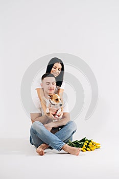 Close up portrait young couple sit and hugging, holding yellow flowers and dog in studio on white background. couple