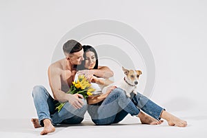 Close up portrait young couple sit and hugging, holding yellow flowers and dog in studio on white background. couple