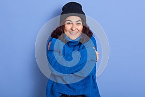 Close up portrait of young curly haired beautiful woman hugging herself, smiling sincerely, looking directly at camera, wearing