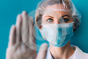 Close-up portrait of young female doctor in medical cap, mask, white gown and gloves on blue background.