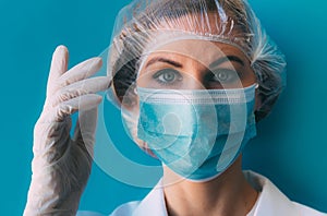 Close-up portrait of young female doctor in medical cap, mask, white gown and gloves on blue background.