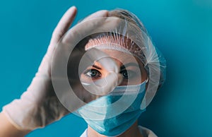 Close-up portrait of young female doctor in medical cap, mask, white gown and gloves on blue background.