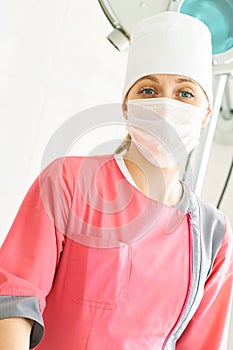 Close-up portrait of young female surgeon doctor in a pink medical gown with a face mask standing in operation theater