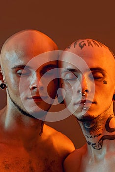 Close up portrait of young half naked twin brothers with tattoos and piercings posing together with eyes closed isolated