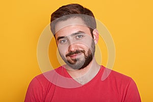 Close up portrait of young handsome man with beard wearing red casual t shirt posing isolated over yellow background, attractive