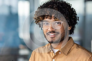 Close-up portrait of young hispanic man wearing glasses, man smiling and looking at camera at workplace inside office