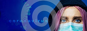 Close-up portrait of young hooded girl with blue eyes and pink hair, wearing medical flu mask with text and icon of coronavirus.