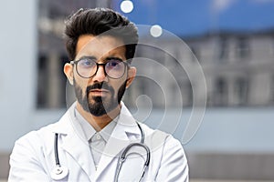 Close up portrait of young Indian doctor, man in white medical coat serious and thinking looking at camera, pediatrician