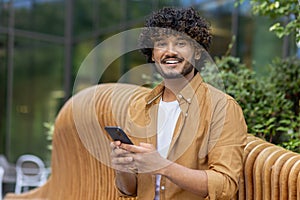 Close-up portrait of a young Indian man sitting outside on a bench, holding a mobile phone and smiling at the camera