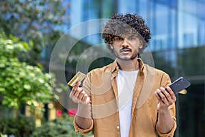 Close-up portrait of a young Indian man standing outside on the street, holding a mobile phone and a credit card