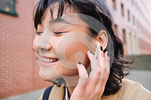 Close up portrait of young korean woman touches earphone, listens music in headphones with pleased smiling face, walks