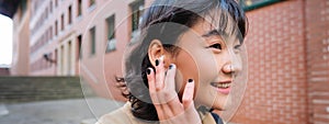 Close up portrait of young korean woman touches earphone, listens music in headphones with pleased smiling face, walks