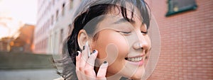 Close up portrait of young korean woman touches earphone, listens music in headphones with pleased smiling face, walks