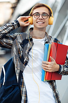 Close-up portrait of young male student in casual outfit with yellow headphones holding folders while listening music on the