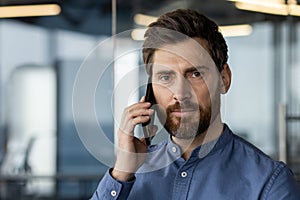 Close-up portrait of a young man in a blue shirt standing in a modern office and talking on the phone, looking