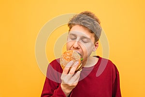 Close-up portrait of a young man in casual clothing bites a burger with his eyes closed and gets pleasure on a yellow background.