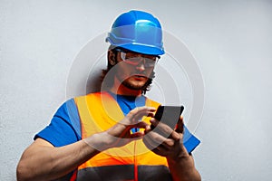 Close-up portrait of young man, construction worker engineer, using smartphone, wearing safety equipment on the grey background.