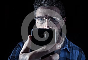 Close up portrait of young man looking intensively to mobile phone screen with blue eyes wide open isolated on black background photo