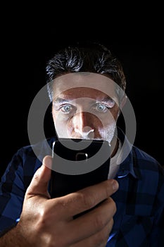 Close up portrait of young man looking intensively to mobile phone screen with blue eyes wide open isolated on black background photo