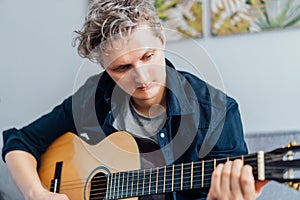 Close up portrait of young man playing acoustic guitar at home sitting on the gray couch, sofa in the living room. New