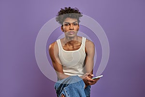 Close-up portrait of young man sits with mobile and looking camera on isolated purple background